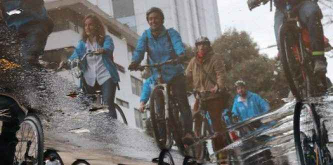 Foto con efecto de reflejo que muestra en un charco de agua en la calle a ciclista usando chaquetas con la imagen de la administración Bogotá Mejor para Todos
